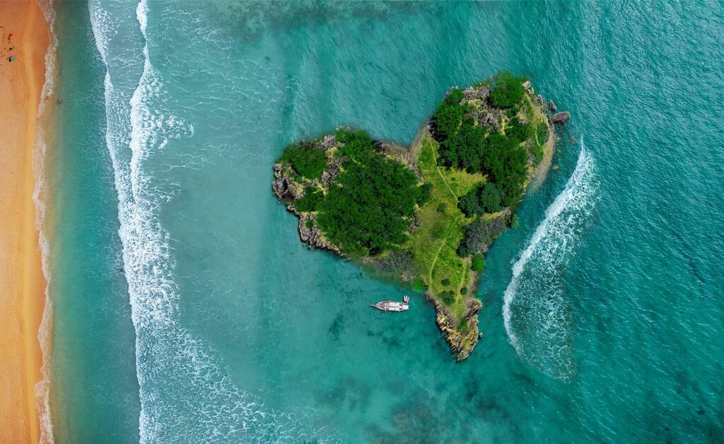 Eine herzförmige, grüne Insel, umgeben von türkisblauem Meer und einem Sandstrand aus der Vogelperspektive.