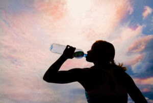 Silhouette einer Frau, die vor einem dramatischen Abendhimmel aus einer Wasserflasche trinkt, um hydriert zu bleiben.