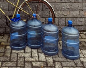 Vier große, blaue Wasserkanister stehen als Trinkwasservorrat aufgereiht auf einem gepflasterten Boden vor einer grauen Steinmauer.