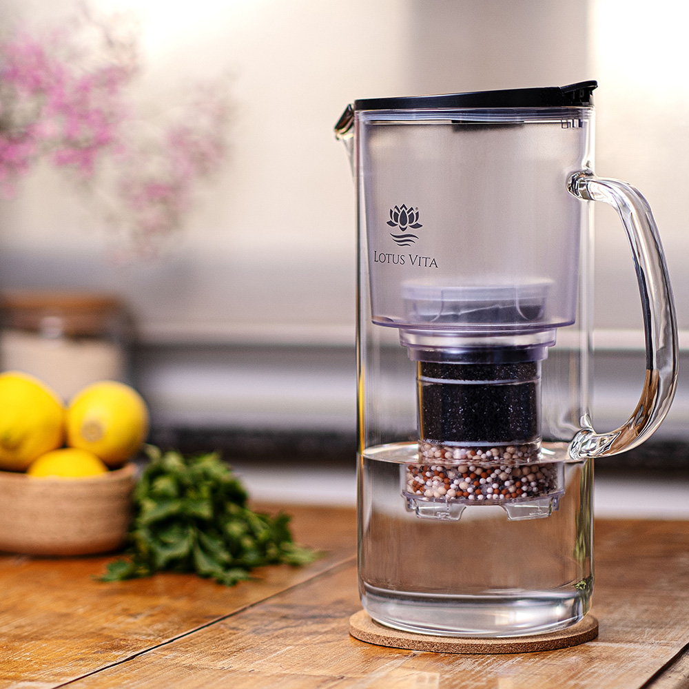 Eine Glas-Wasserfilterkanne von Lotus Vita steht auf einem Holztisch, gefüllt mit gefiltertem Wasser, umgeben von frischen Zitronen und Minze.