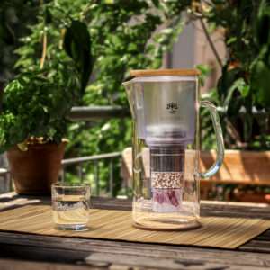 Eine Glaskaraffe mit integriertem Wasserfilter und Bambusdeckel steht neben einem gefüllten Wasserglas auf einem Holztisch auf einem Balkon.