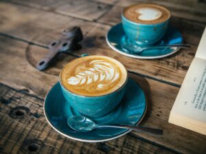 Zwei türkisfarbene Tassen Cappuccino mit Latte Art stehen auf einem rustikalen Holztisch neben einem aufgeschlagenen Buch und schaffen eine gemütliche Atmosphäre.