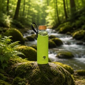 Eine Glas-Trinkflasche mit hellgrüner Schutzhülle und Holzdeckel steht auf einem moosbewachsenen Stein an einem Bach im Wald.