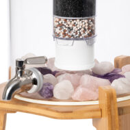 Ein Glas-Wasserfilter mit mehrstufiger Filterkartusche und Edelstahl-Zapfhahn, auf einem Bett aus Rosenquarz- und Amethyst-Edelsteinen in einem Behälter auf einem Holzständer.